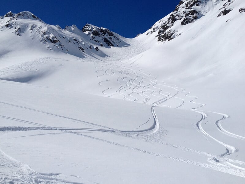 Pente enneigée avec traces de ski en courbes, entourée de montagnes rocheuses sous un ciel clair.