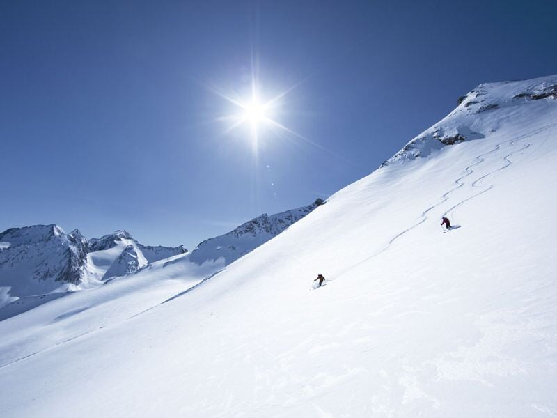 Deux skieurs dévalant une pente enneigée sous un soleil éclatant et un ciel bleu sans nuages.