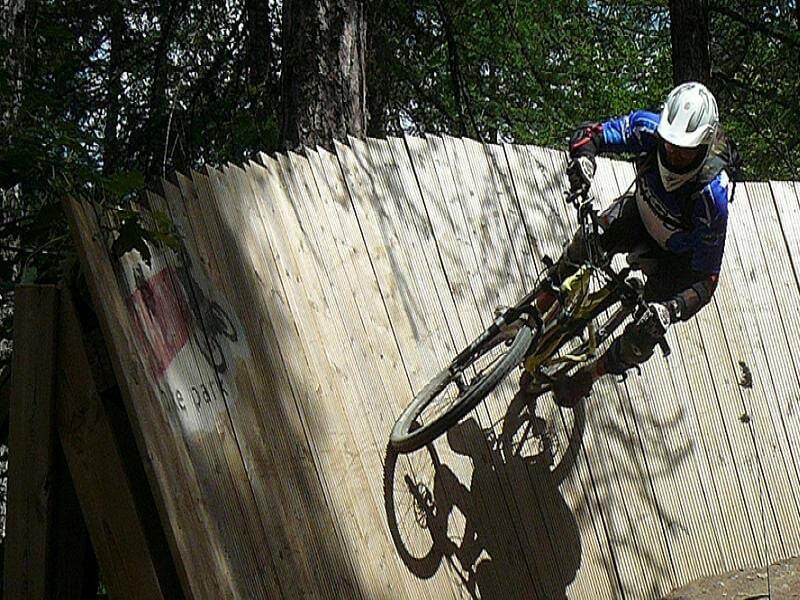 Cycliste en tenue de protection roulant sur une piste en bois dans une forêt dense et ombragée.
