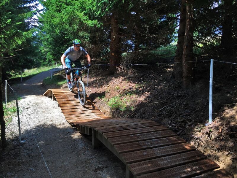 Cycliste en tenue sportive franchissant un pont en bois sur un sentier forestier en montagne.