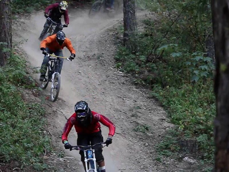 Trois vététistes en tenue de protection descendant un sentier étroit et poussiéreux en forêt.