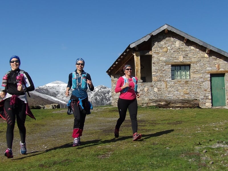 Trois femmes courant sur un chemin de montagne près d'une maison en pierre avec des montagnes enneigées à l'arrière-plan.