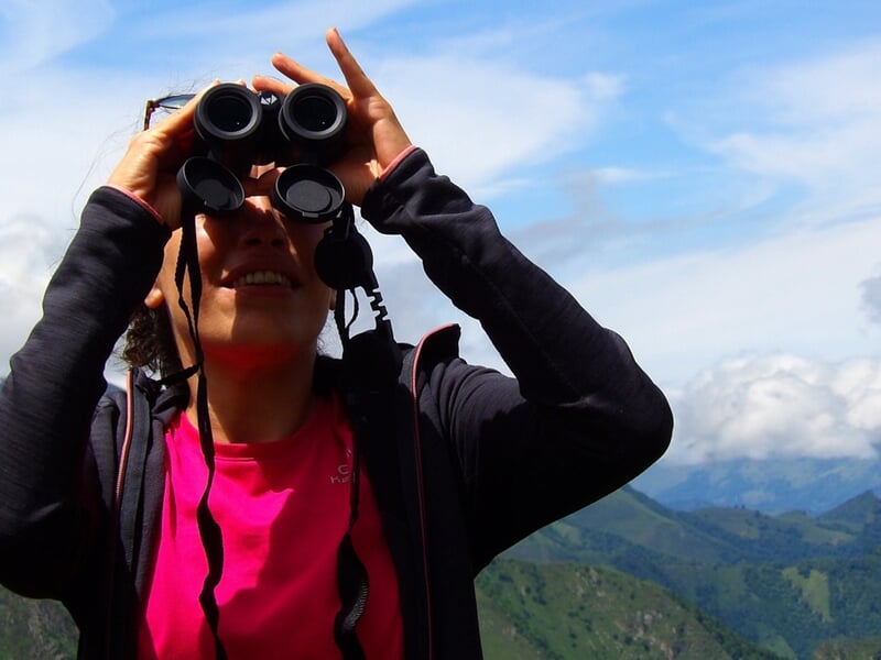 Femme regardant au loin à travers des jumelles, en pleine nature avec des montagnes en arrière-plan.