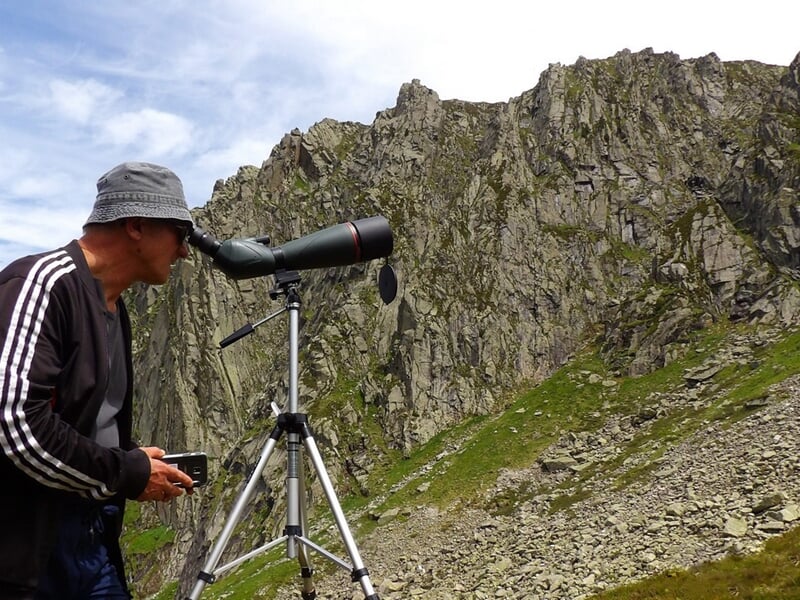 Homme utilisant une longue-vue sur trépied pour observer la nature dans un paysage montagneux rocheux.