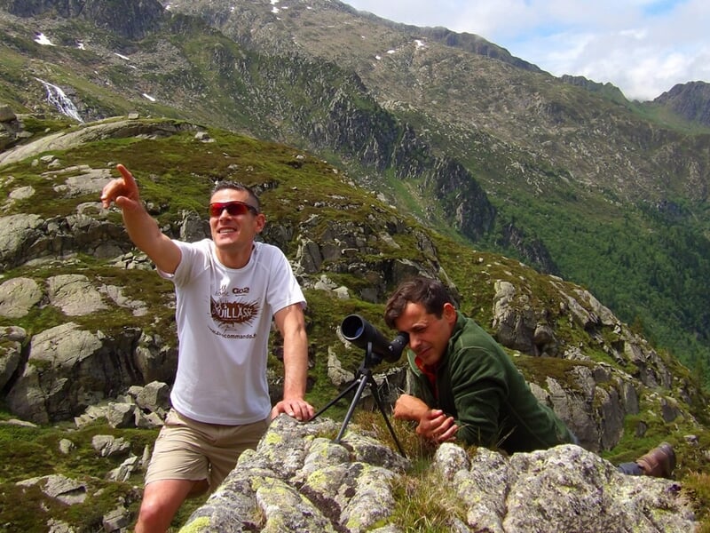 Deux hommes en montagne, l'un pointant du doigt, l'autre regardant à travers une longue-vue posée sur un rocher.