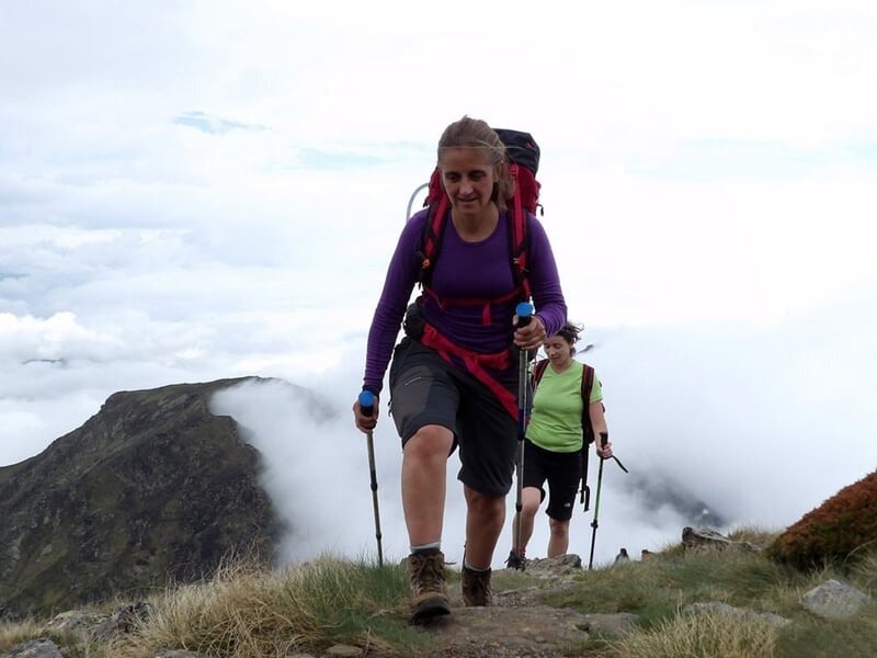 Deux randonneuses en montagne, l'une au premier plan souriante, l'autre en arrière-plan sur un sentier.