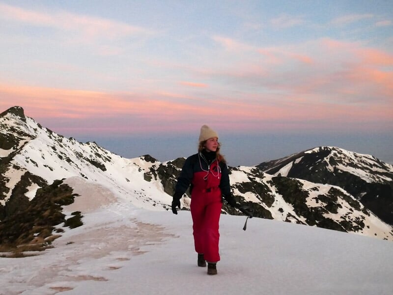 Femme marchant seule sur un sentier enneigé au coucher du soleil, entourée de montagnes.