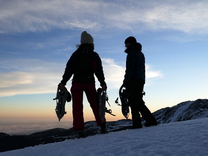 Deux personnes debout sur la neige au crépuscule, équipées de raquettes à neige, silhouette contre le ciel.