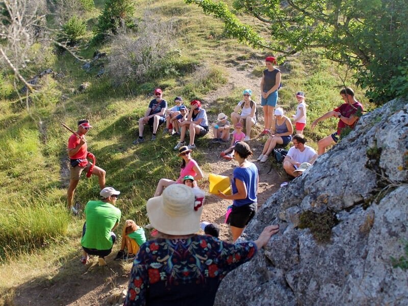 Groupe de randonneurs assis et écoutant un guide en pleine nature, sur un terrain herbeux et rocheux.