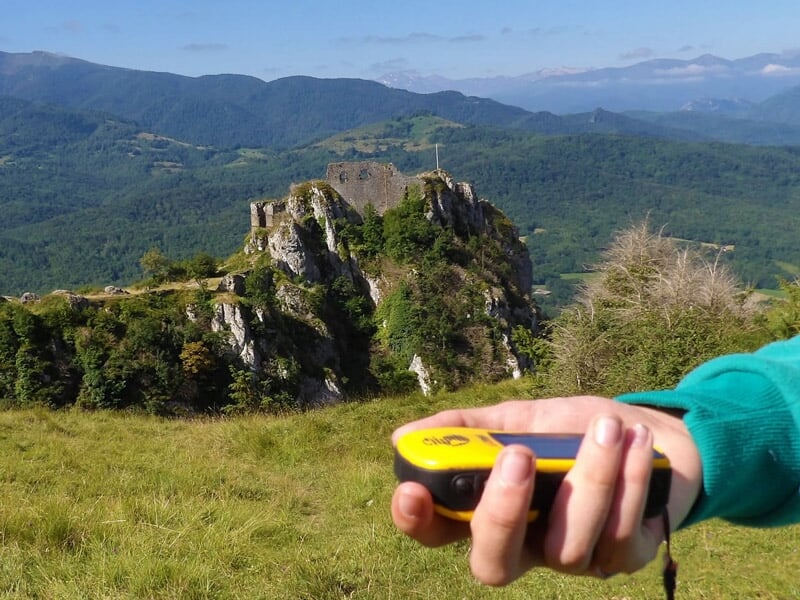 Vue d'un château en ruines sur une colline verdoyante, avec une main tenant un GPS au premier plan.