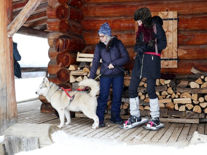 Deux personnes avec un chien de traîneau Husky se tenant à côté d'un abri en bois dans un cadre enneigé