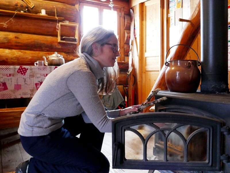 Femme alimentant un poêle à bois dans une cabane rustique avec une marmite suspendue au-dessus