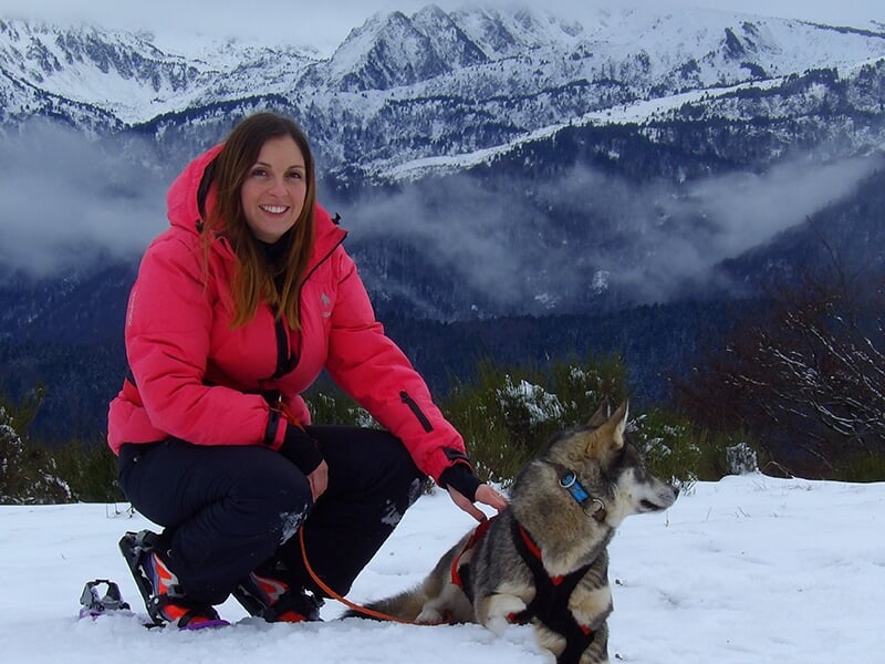 Une femme portant une veste rose dans un paysage de montagne enneigé, posant avec un husky.