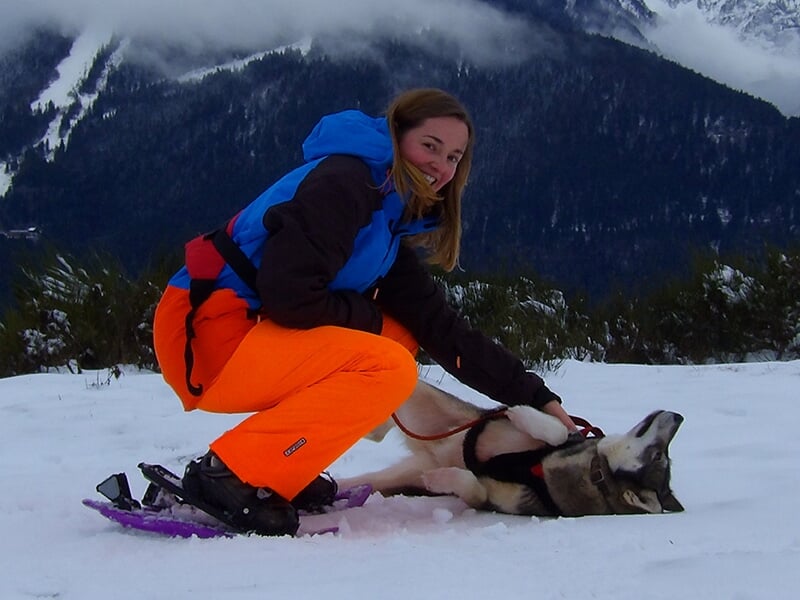 Une femme en tenue d'hiver jouant avec un husky couché dans la neige, entourée de montagnes.