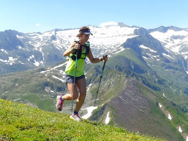 Femme courant sur un sentier de crête avec des bâtons de randonnée, entourée de montagnes.