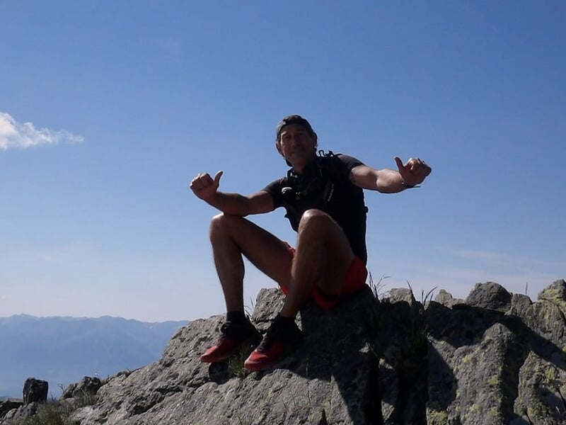 Homme assis sur un sommet rocheux, levant les bras dans une posture de victoire avec des montagnes à l'arrière-plan.