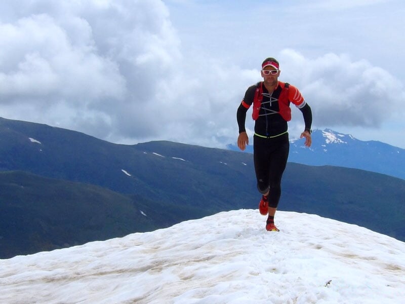 Trail runner portant une tenue noire et rouge, courant sur une crête enneigée avec un panorama de montagnes et ciel nuageux