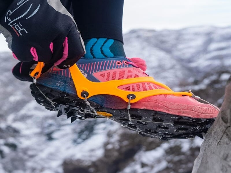 Chaussure de trail rose équipée de crampons antidérapants orange, en train d’être ajustée par une main gantée. Décor montagnard enneigé en arrière-plan
