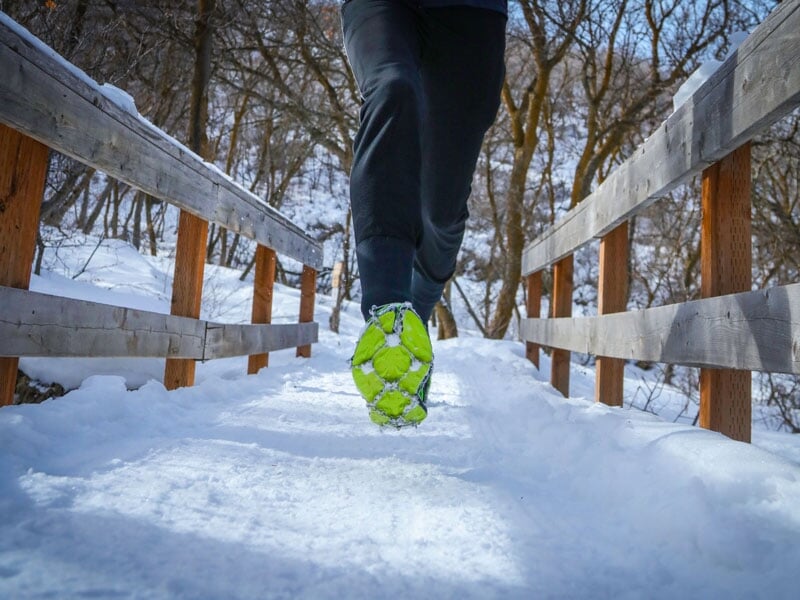 Gros plan sur des chaussures de trail avec semelles vertes cramponnées, en mouvement sur un pont en bois recouvert de neige, entouré d’arbres dénudés en hiver