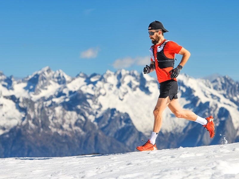 Homme courant sur un sentier enneigé en haute montagne, vêtu de rouge et noir, avec des chaussures de trail running rouges. Chaîne de montagnes aux sommets enneigés en arrière-plan sous un ciel bleu