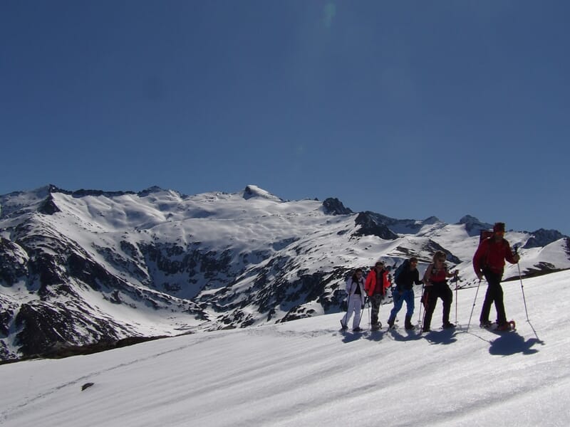 Cinq randonneurs en raquettes à neige progressant sur un versant enneigé avec montagnes en arrière-plan sous un ciel clair.