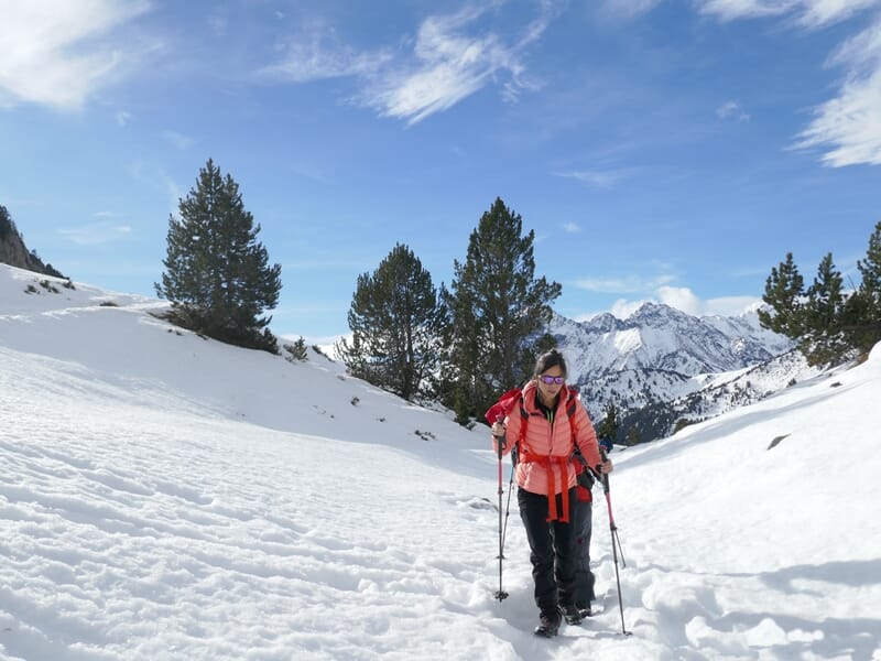 Femme en tenue rouge et noire marchant en raquettes à neige dans un paysage montagneux enneigé avec arbres et ciel bleu.