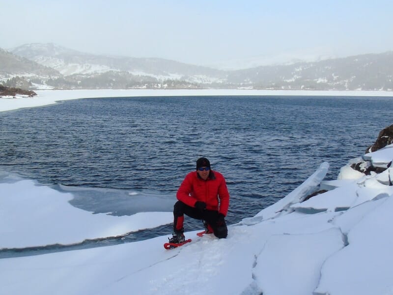 Personne en raquettes à neige près d'un lac partiellement gelé entouré de montagnes enneigées sous un ciel clair.