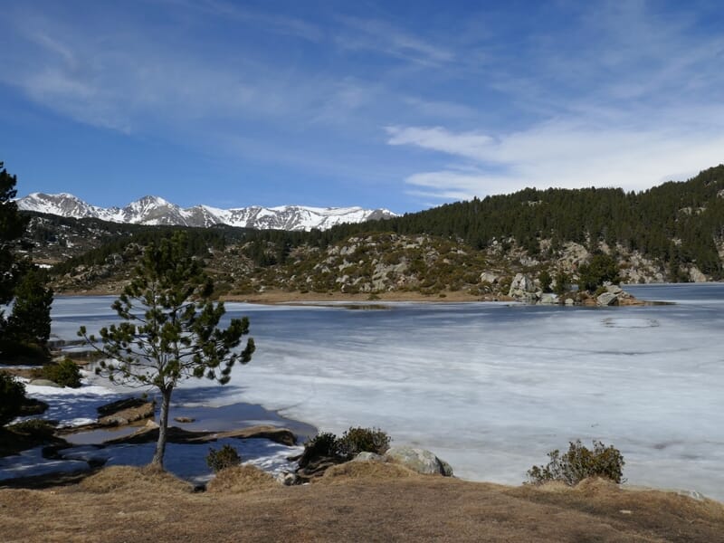 Lac gelé avec des arbres et des rochers au bord, montagnes enneigées au loin sous un ciel bleu avec quelques nuages.