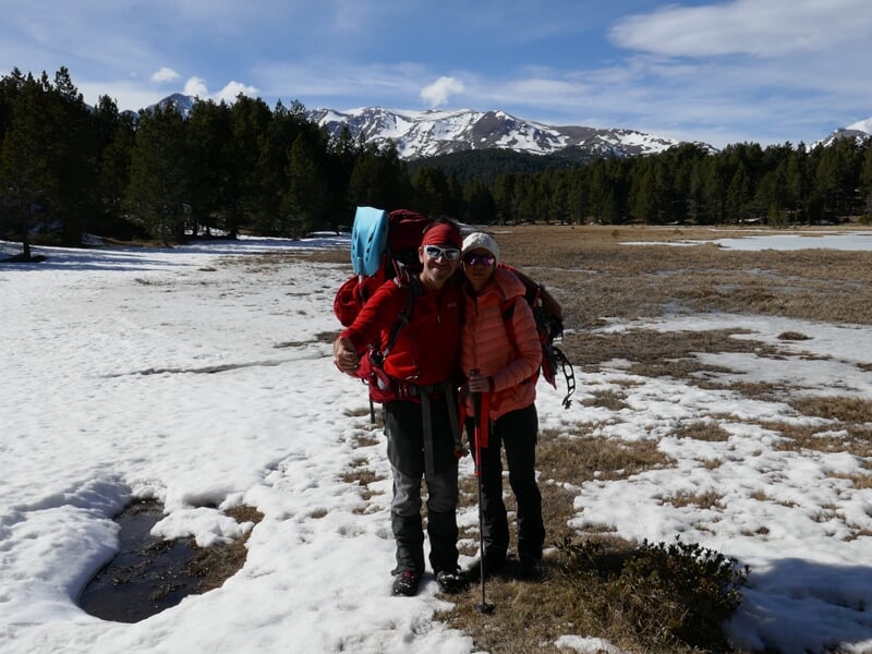 Couple en tenue de randonnée avec sacs à dos et bâtons, debout sur un sol partiellement enneigé en montagne.