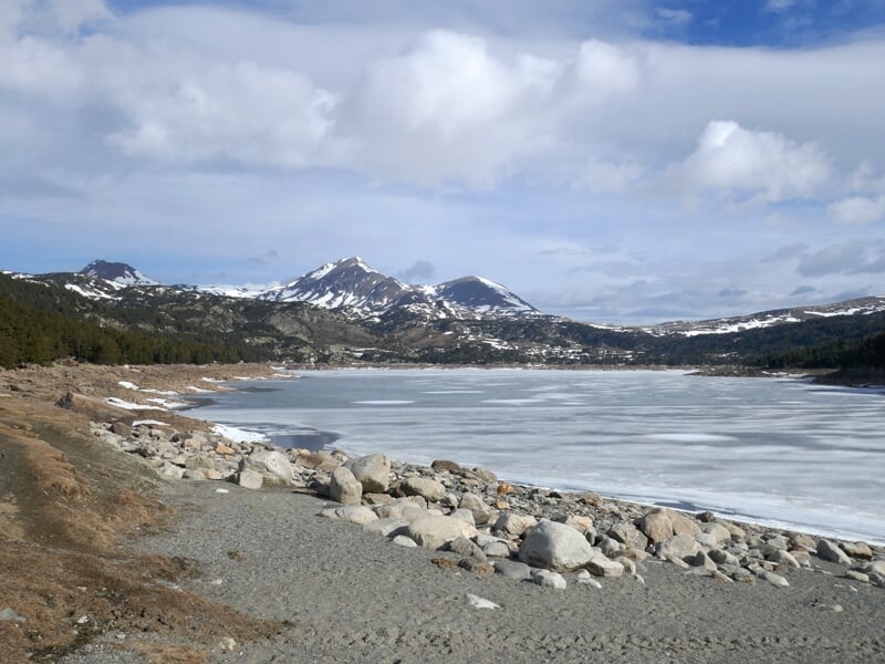Vue d'un lac gelé bordé de rochers, avec des montagnes enneigées et une forêt dense sous un ciel nuageux.