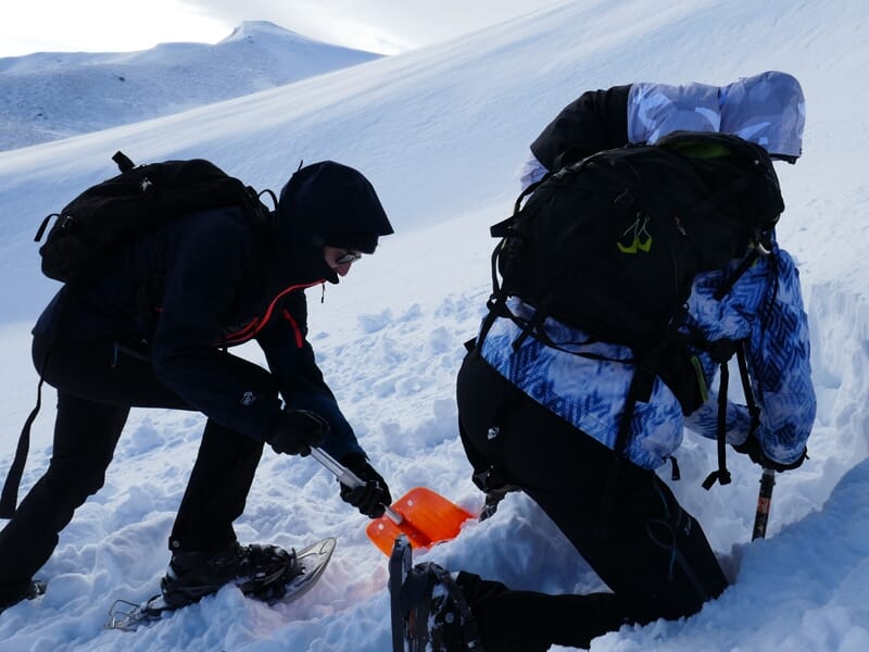 Deux personnes en tenue d'hiver fouillent la neige avec une pelle orange dans une station de ski enneigée.
