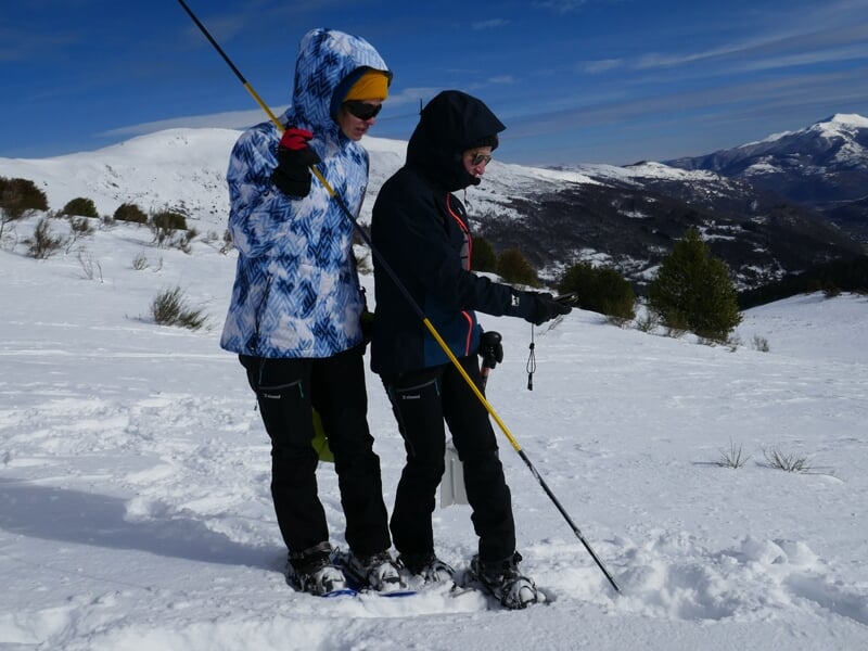 Deux personnes en vêtements chauds utilisent un détecteur et une sonde dans la neige en montagne sous un ciel bleu.