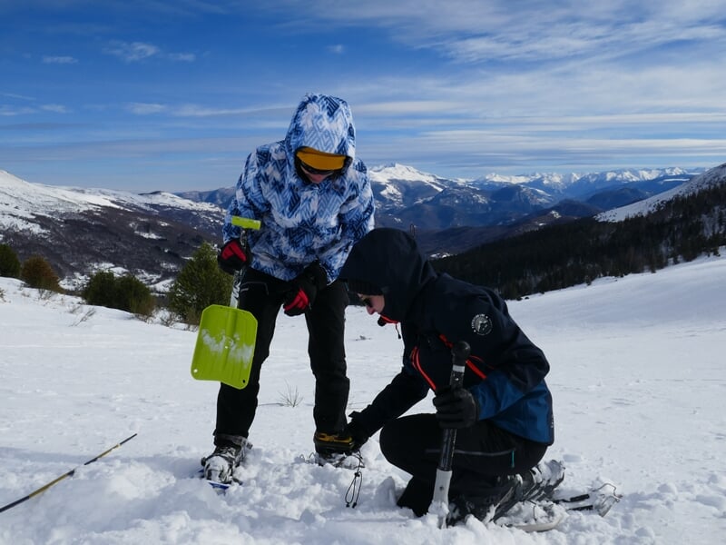 Deux personnes en tenue d'hiver examinent la neige avec une pelle et une sonde dans un paysage montagneux enneigé.