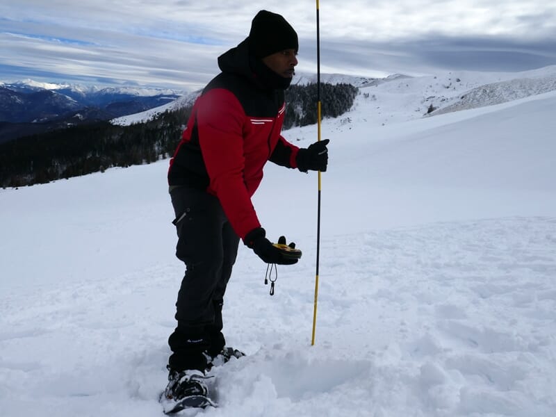 Homme en tenue rouge et noire utilise une sonde pour sonder la neige dans une station de ski en montagne.