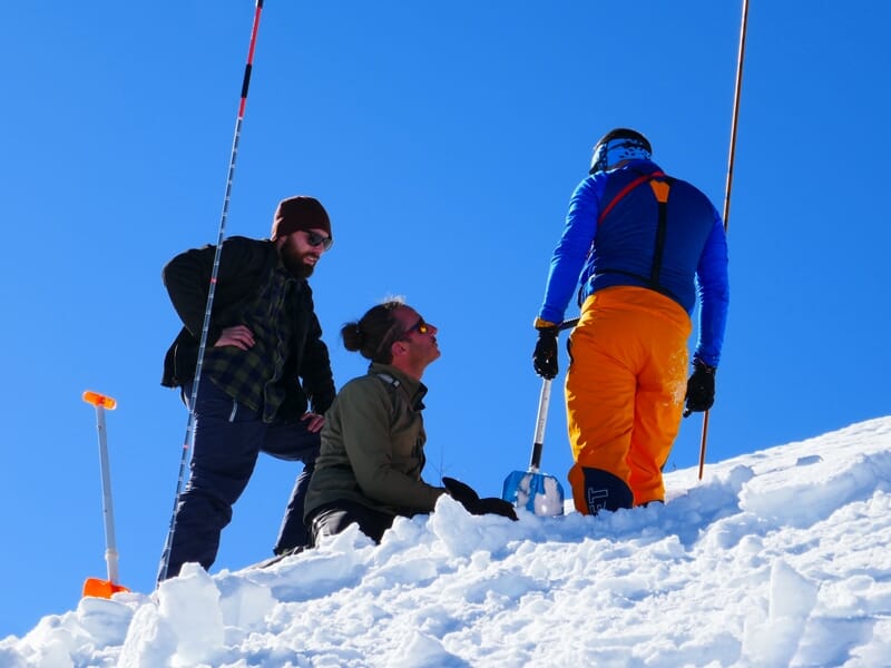 Trois personnes en tenue de ski discutent sur une crête enneigée avec un ciel bleu et montagnes en arrière-plan.