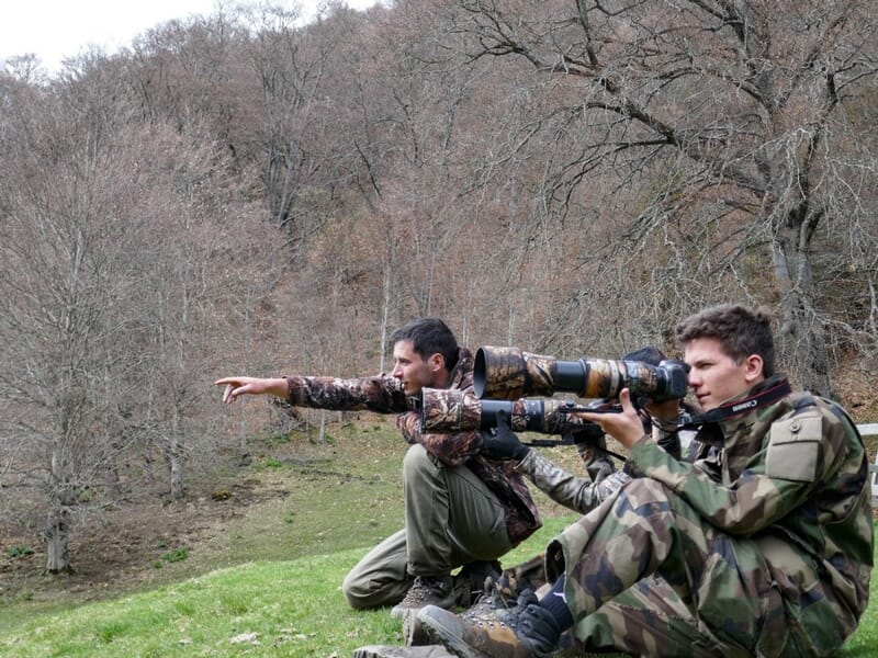 Deux hommes en tenue camouflage observant la nature, l'un pointant du doigt, l'autre avec un appareil photo à longue focale.