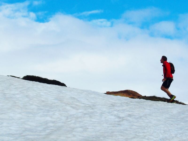 Coureur en montée sur un champ de neige, vêtu d’un haut rouge et portant un petit sac à dos, avec un ciel partiellement nuageux