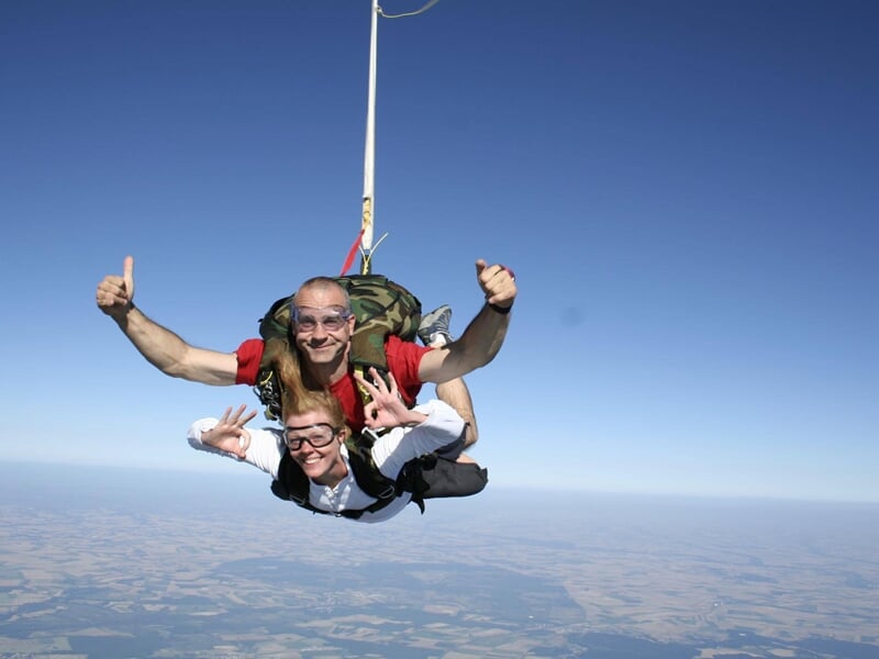 Deux personnes en saut en parachute en tandem, souriantes, avec le pouce levé et ciel bleu dégagé en arrière-plan.