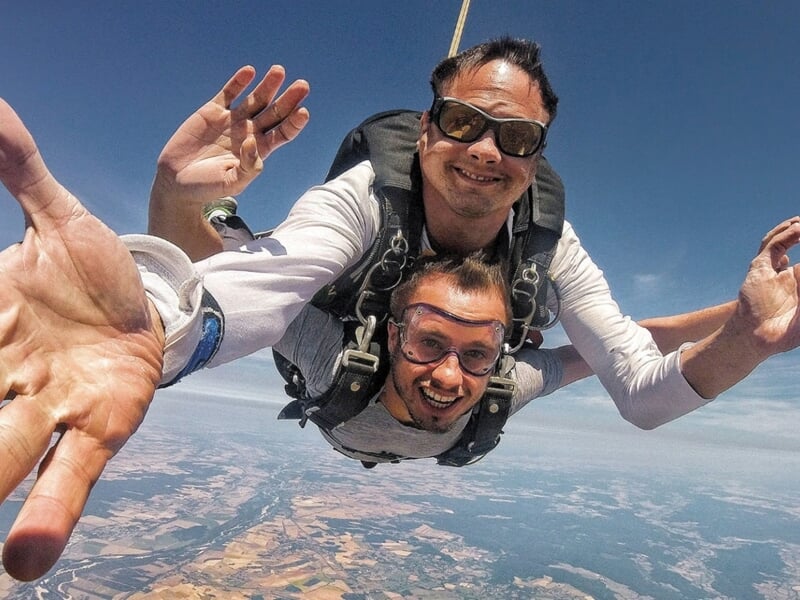 Saut en parachute en tandem avec deux hommes souriants, l'un tendant la main vers la caméra, ciel bleu clair.