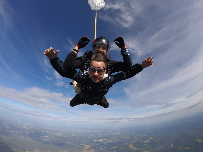 Saut en parachute en tandem avec deux hommes en combinaison noire, ciel nuageux et paysage visible en dessous.