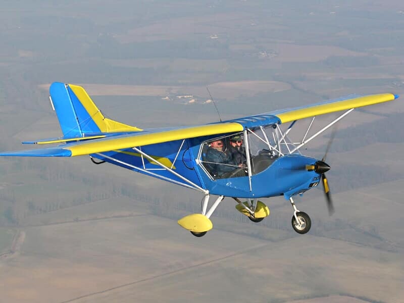 Un avion ultraléger bleu et jaune vole dans le ciel avec deux personnes visibles dans le cockpit.