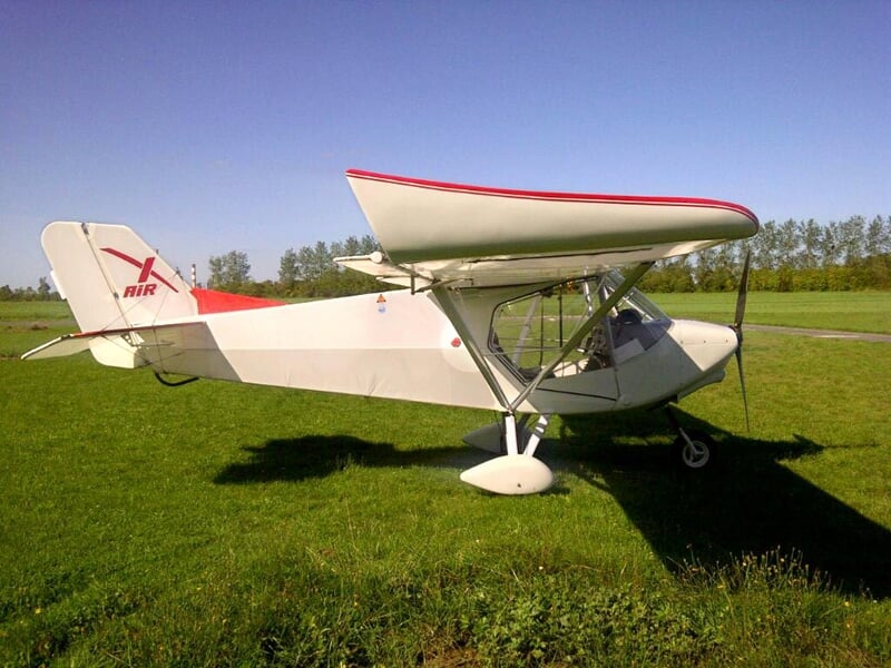 Un avion ultraléger blanc avec des détails rouges est stationné sur une pelouse verte sous un ciel clair.