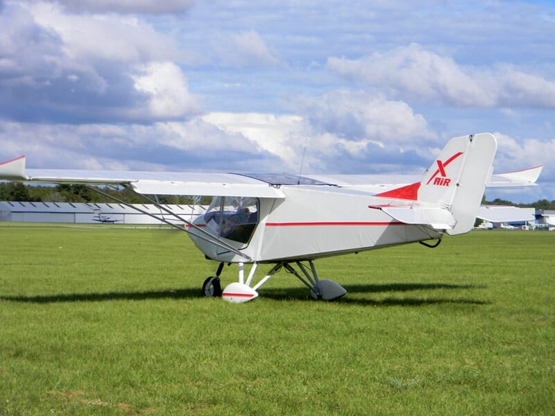 Un avion ultraléger blanc avec des marques rouges est posé sur une grande étendue d'herbe verte sous un ciel nuageux.