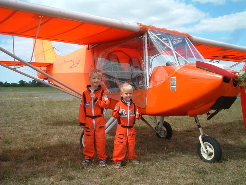 Deux enfants en combinaison orange posent devant un petit avion ultraléger orange sur un terrain d'herbe.
