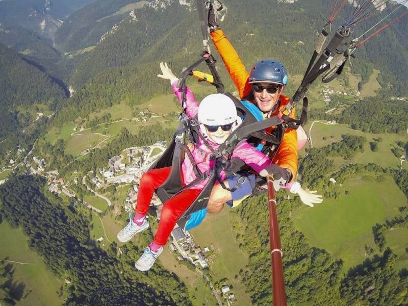 Personne en tandem parapente avec instructeur souriant, survolant un village et des montagnes verdoyantes.