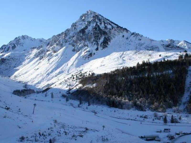 Montagne enneigée avec un ciel bleu clair, forêt dense au pied et pistes de ski visibles en bas.