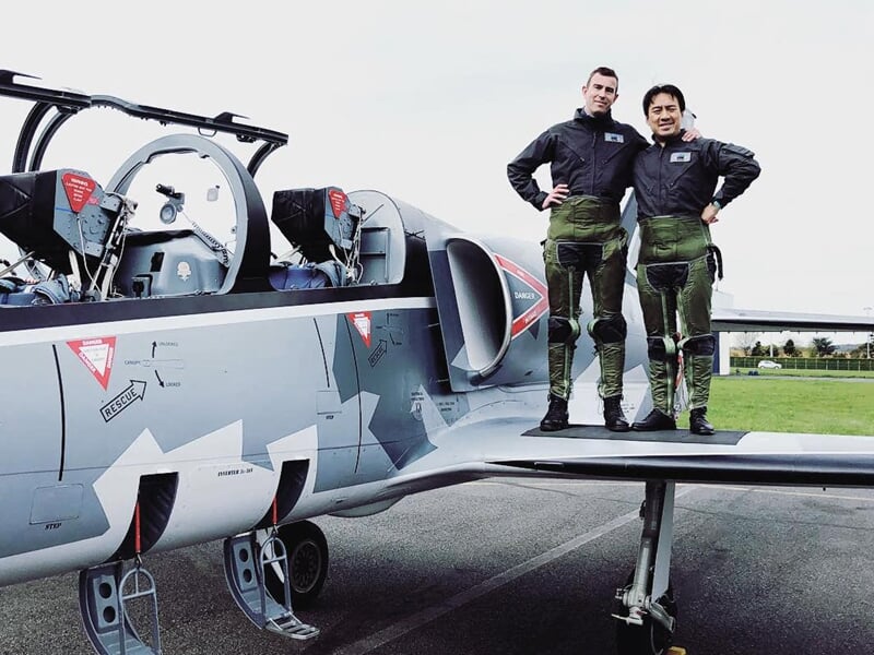 Deux pilotes sur l'aile d’un L-39 Albatros au sol, livrée camouflage avec marquages rouges.