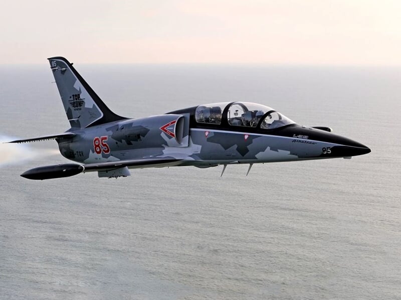 L-39 Albatros en vol au-dessus de la mer, livrée camouflage gris et noir, numéro 85 rouge sur le fuselage.