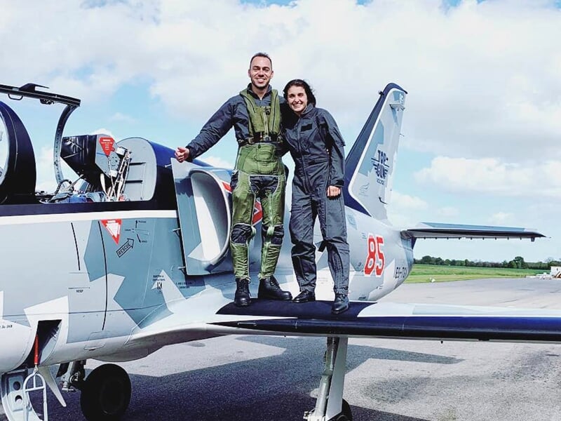 Deux pilotes souriants en combinaison debout sur l'aile d'un avion de chasse camouflé sur la piste.