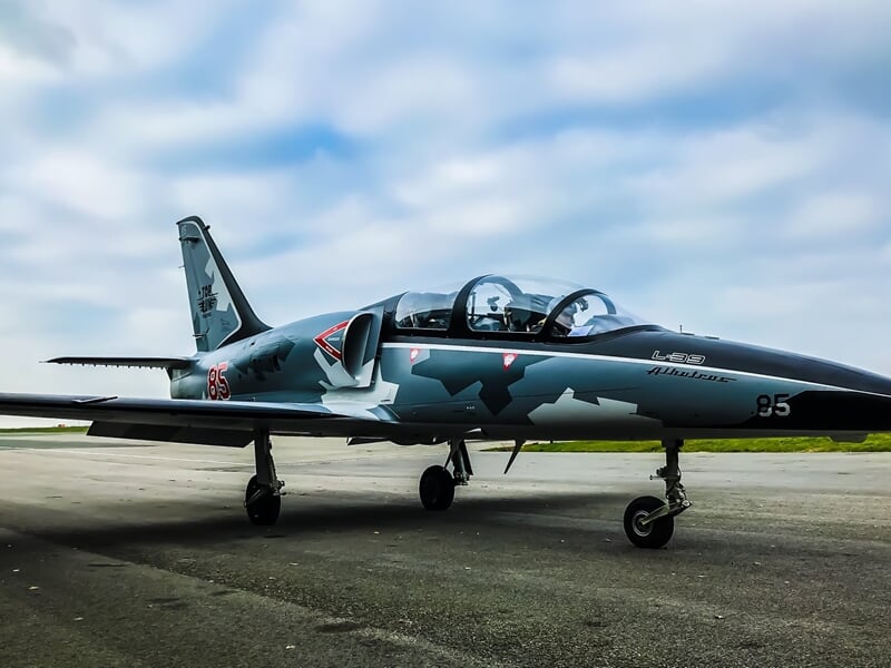 Avion de chasse camouflé gris stationné sur une piste d'aéroport sous un ciel nuageux.